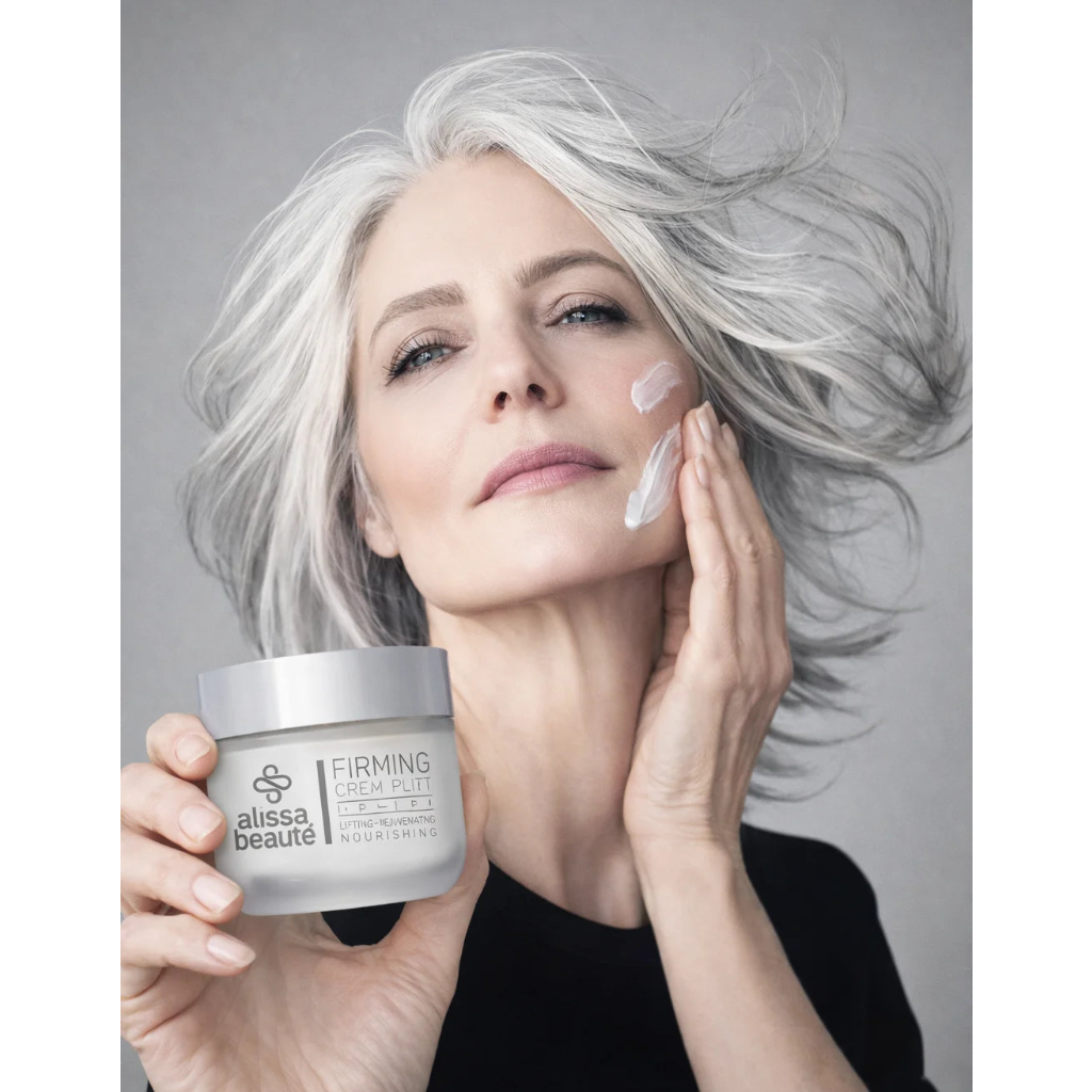 Femeie elegantă cu păr argintiu în mișcare aplicând Firming Cream Alissa Beauté pe obraz cu mișcări ascendente, borcan 50ml vizibil în mâna stângă cu inscripția Lifting – Rejuvenating – Nourishing. Ten matur ferm și luminos, fond gri, top negru. Cremă anti-îmbătrânire cu Biollagen colagen vegan și Bio GF Complex, linia UPLIFT.
