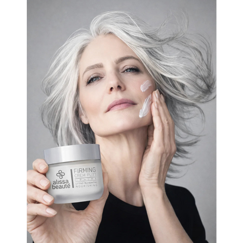 Femeie elegantă cu păr argintiu în mișcare aplicând Firming Cream Alissa Beauté pe obraz cu mișcări ascendente, borcan 50ml vizibil în mâna stângă cu inscripția Lifting – Rejuvenating – Nourishing. Ten matur ferm și luminos, fond gri, top negru. Cremă anti-îmbătrânire cu Biollagen colagen vegan și Bio GF Complex, linia UPLIFT.