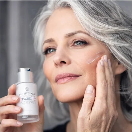 Femeie cu ten matur aplicând Firming Booster Alissa Beauté pe pomeți, flacon gri cu pompă ținut în mână stângă. Booster anti-rid cu exosomi AC ExoTone și efect blurring vizibil imediat, linia UPLIFT.
