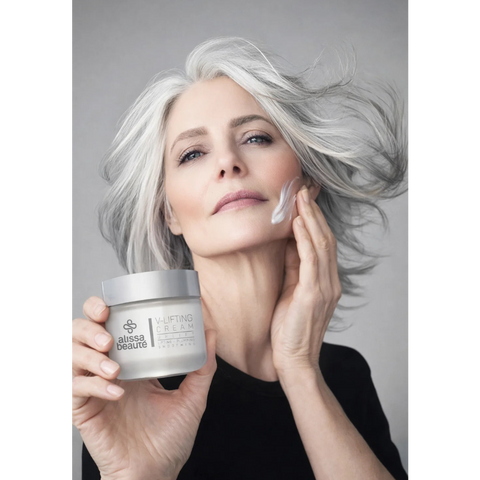 Femeie elegantă cu păr argintiu în mișcare aplicând V-Lifting Cream Alissa Beauté pe obraz cu mișcări ascendente, borcan vizibil în mâna stângă. Ten matur ferm și radiant, fond gri, top negru. Cremă lifting cu Centella Reversa și Biollagen colagen vegan, linia UPLIFT.